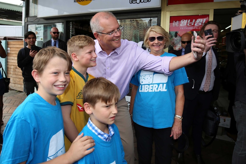 Scott Morrison smiles for a selfie with three children. A woman in a Liberal shirt is smiling at the ABC's photographer