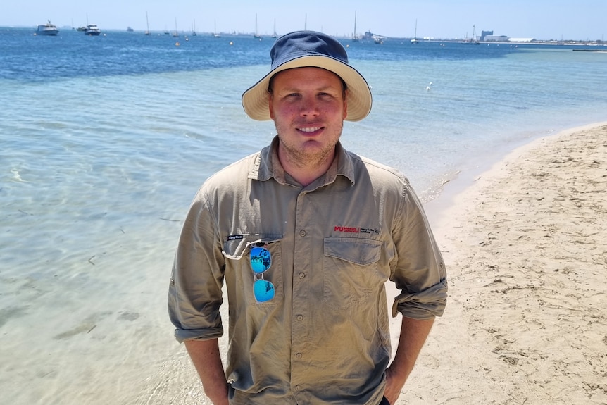 man standing with blue wide brimmed hat on on the beach with blue water behind him and the sun shining