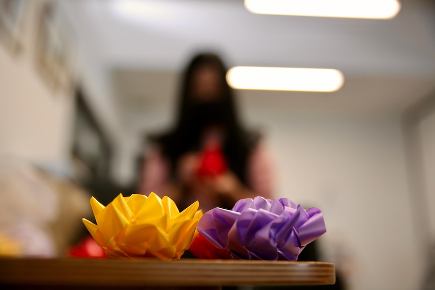 An image focusing on colourful flowers made of ribbons in the foreground and the blurred image of a woman in the background.