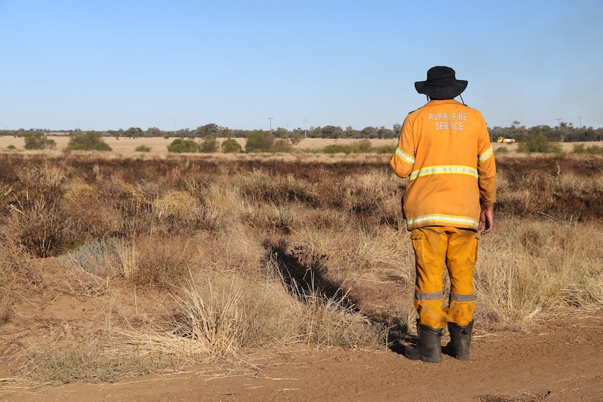 A man in firefighter gear stands on the side of a dirt road, looking out over a bone-dry landscape.