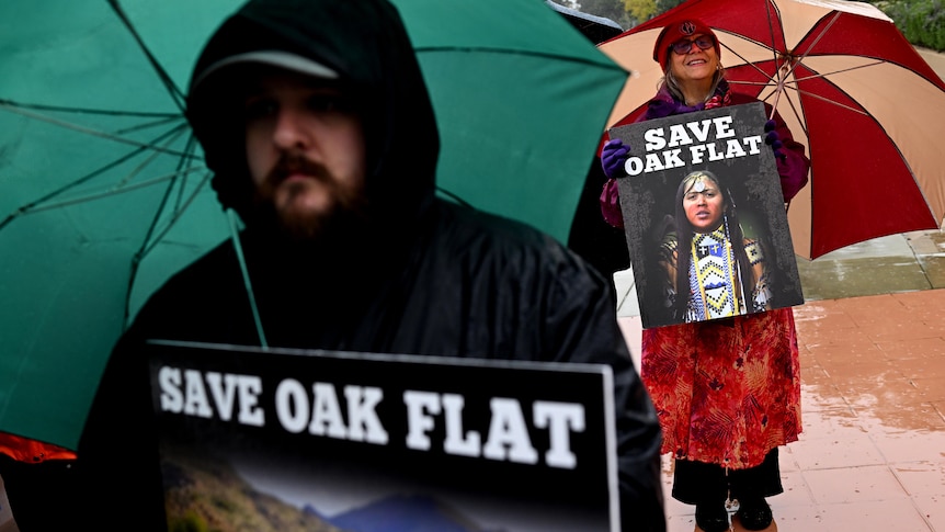 A man and woman in raincoats hold umbrellas and signs saying 'save Oak Flat'.