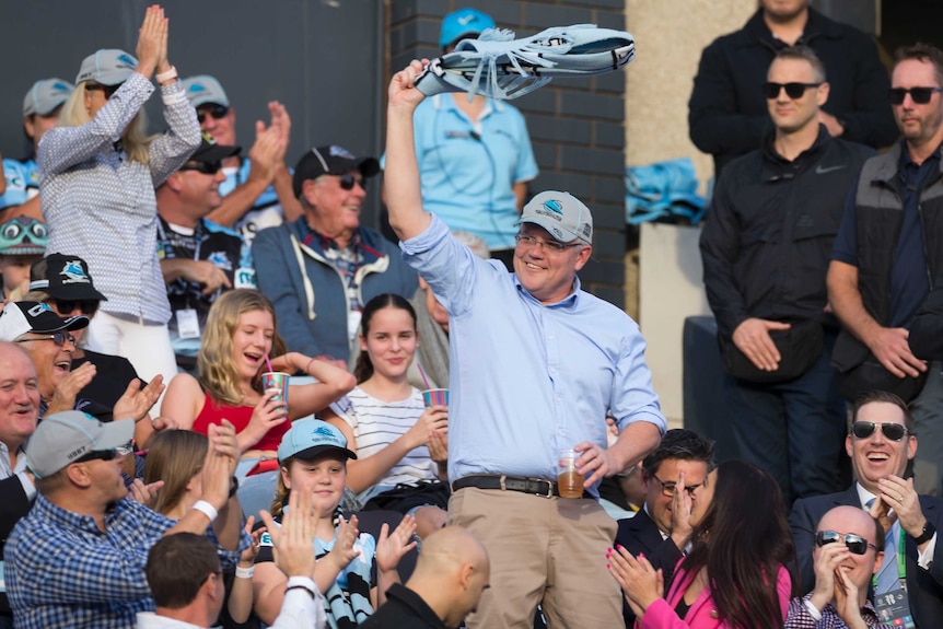 Scott Morrison in a grandstand swinging a Sharks jersey around his head, holding a beer.