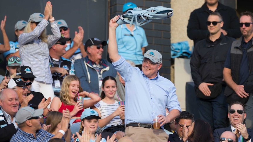 Scott Morrison in a grandstand swinging a Sharks jersey around his head, holding a beer.