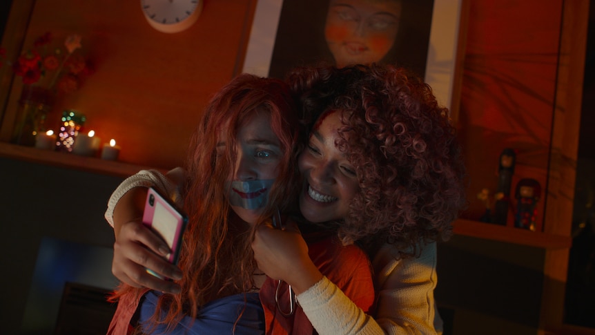 A young woman of colour smiles and holds a phone out to a blonde white woman who is tied to a chair with tape around her mouth.