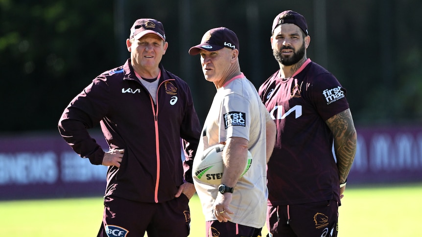 An NRL coach, trainer and player stand on a field watching training.