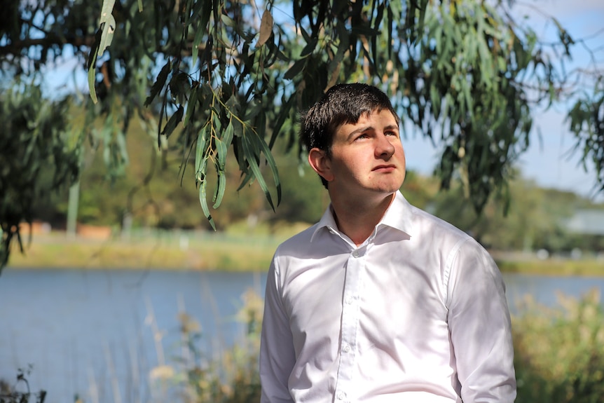 Young man wearing white shirt stands in dappled light beneath tree with river and grassy banks in the backgrounf