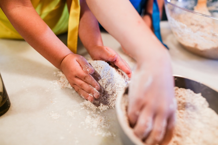 A child's hands gather up flour on a bench, while an adult hand reaches into a mixing bowl