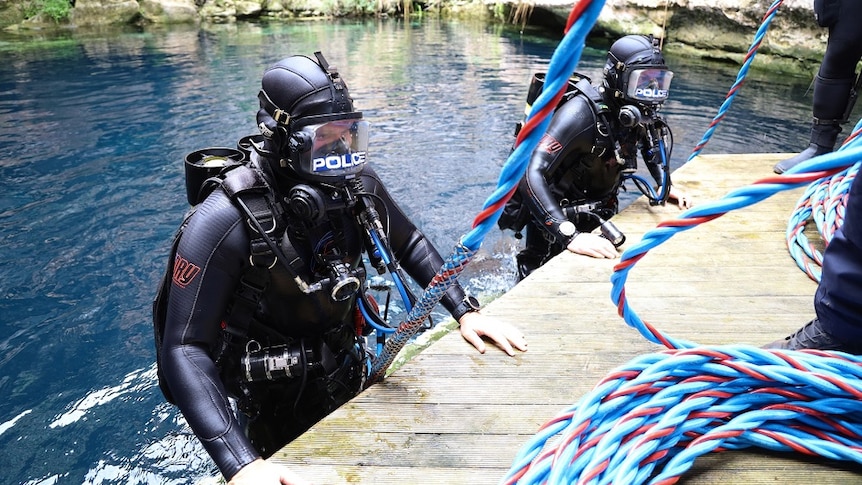 Two divers in wetsuits and masks get out of the water onto a platform. Blue and red spired ropes are connected to them.