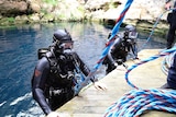 Two divers in wetsuits and masks get out of the water onto a platform. Blue and red spired ropes are connected to them.