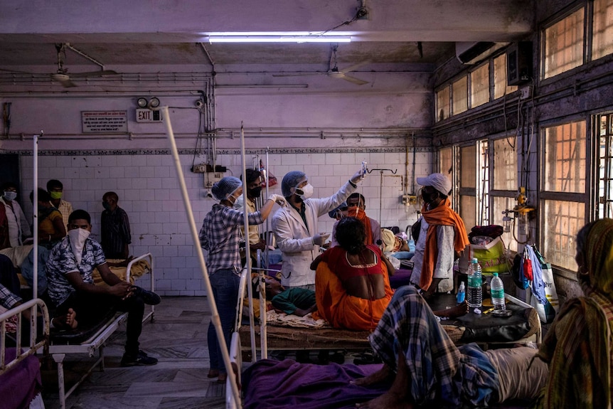 A man wearing scrubs and a mask holds an IV as men sit on hospital beds around him in a run down hospital room.