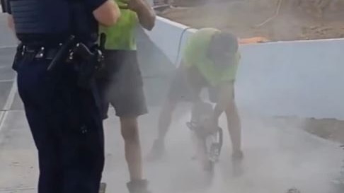 A tradie in hi-vis jackhammers a concrete driveway as another tradie speaks with a policeman
