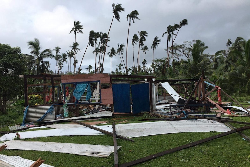 A flattened tin building under a grey sky, with coconut trees behind it.