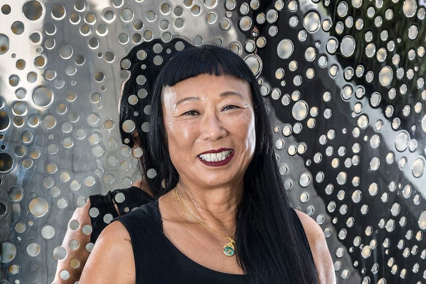 portrait of Asian woman wearing black dress and smiling. Behind her is a silver metal artwork with many holes
