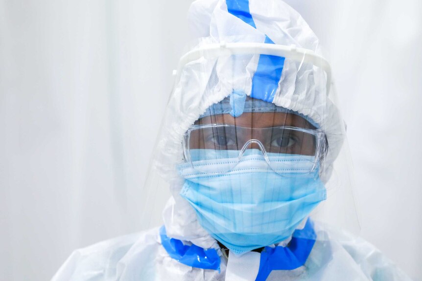 A woman in full PPE stands against a white curtain