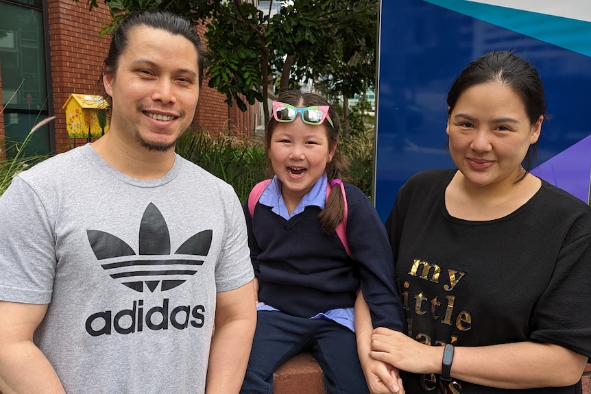 A man, standing next to a young child wearing a school uniform who is next to a woman who is her mother.