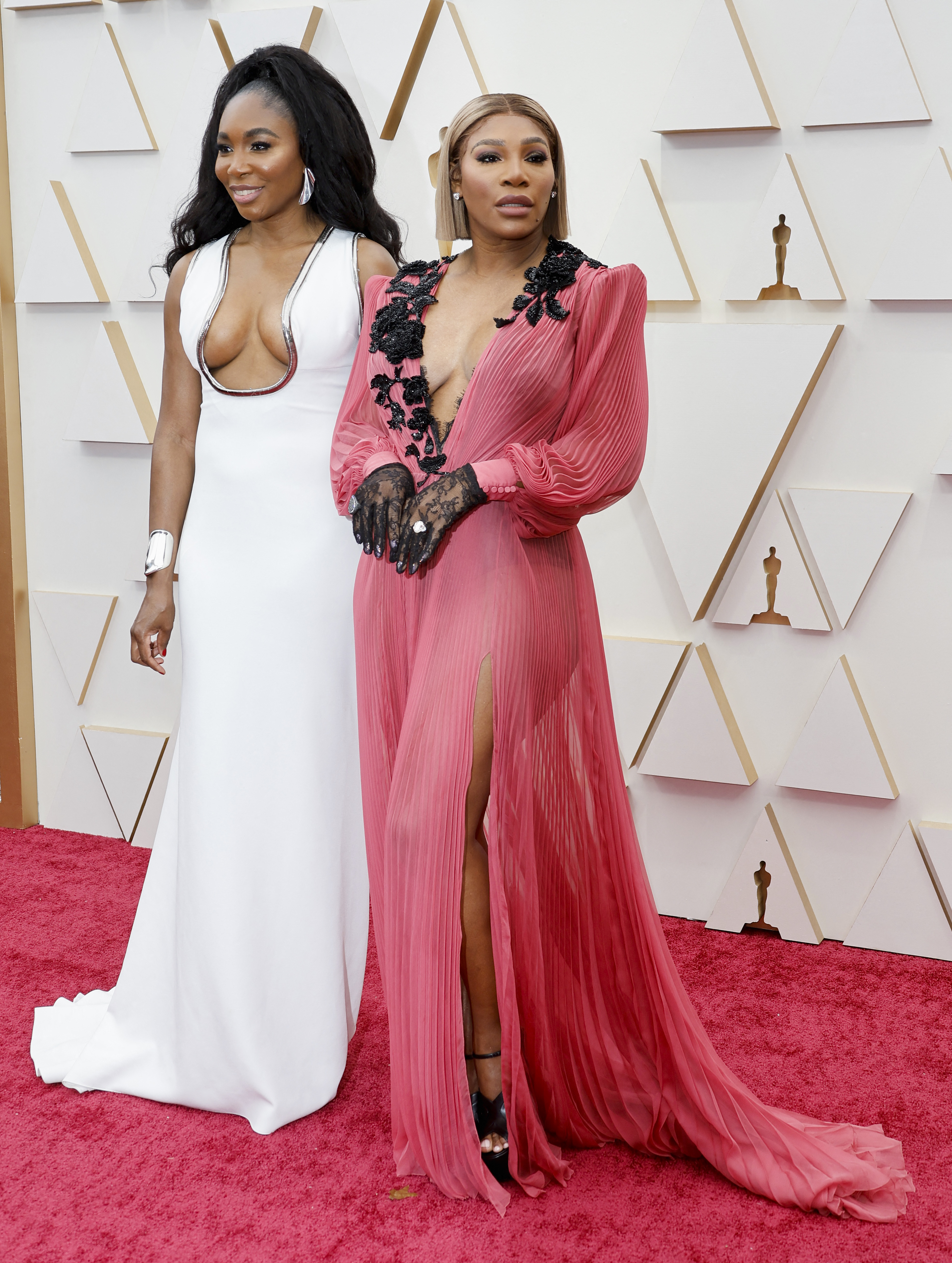 tennis players venus and serena williams pose on the red carpet. venus wears a white plunging gown, serena wears pink flowing 
