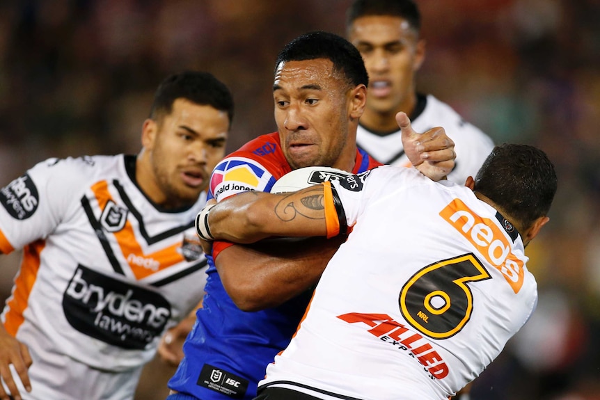 A male NRL player holds the ball with his right arm across his chest as he is tackled around the arms.