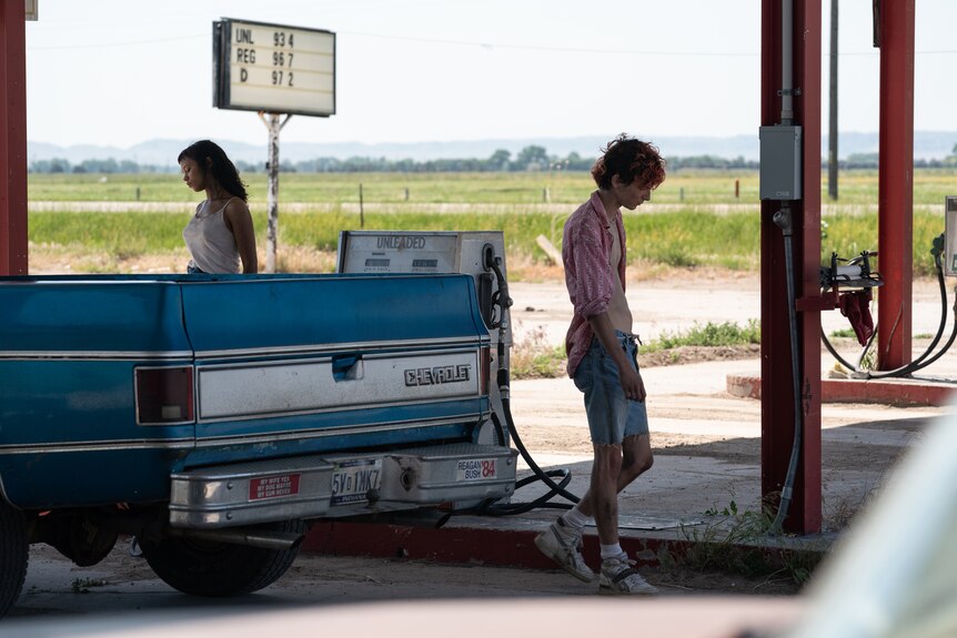 A young Black woman and a pale young man with dyed red hair stand near a blue truck at a petrol station