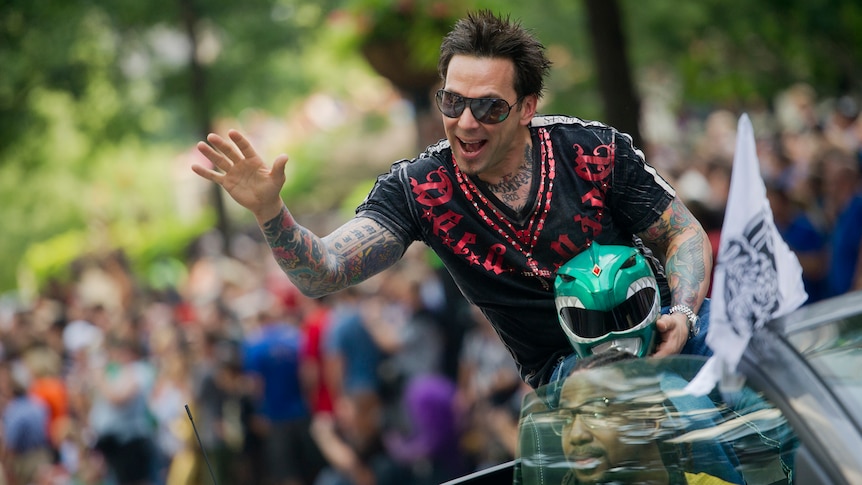 A man is pictured in a convertible while waving at a large crowd. In his hands the Green Power Ranger helmet.