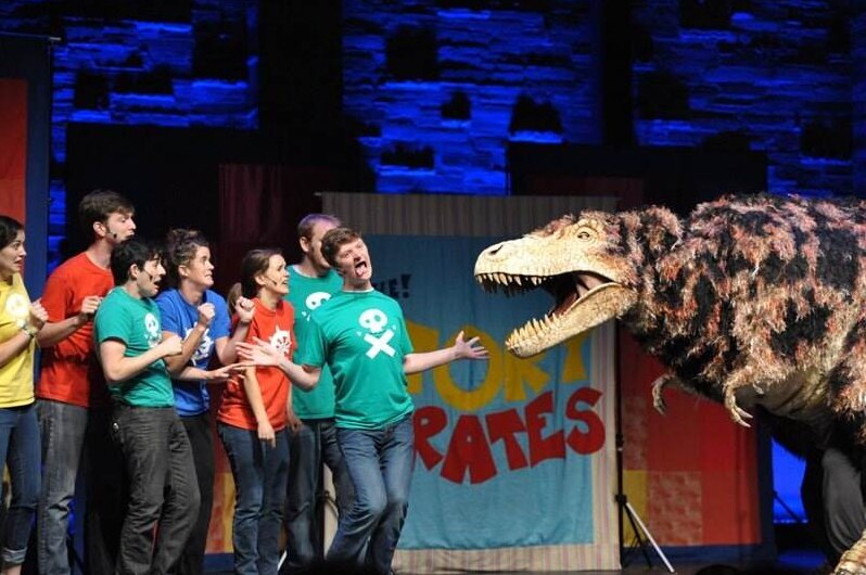 A group of actors from the podcast Story Pirates on stage with a dinosaur.