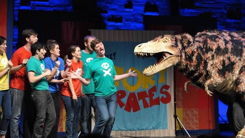 A group of actors from the podcast Story Pirates on stage with a dinosaur.