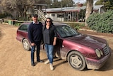 A couple dressed in casual clothes stands in front of a maroon Mercedes Benz wagon. 