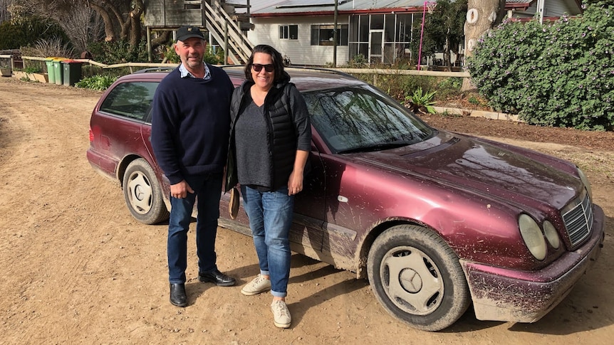 A couple dressed in casual clothes stands in front of a maroon Mercedes Benz wagon. 