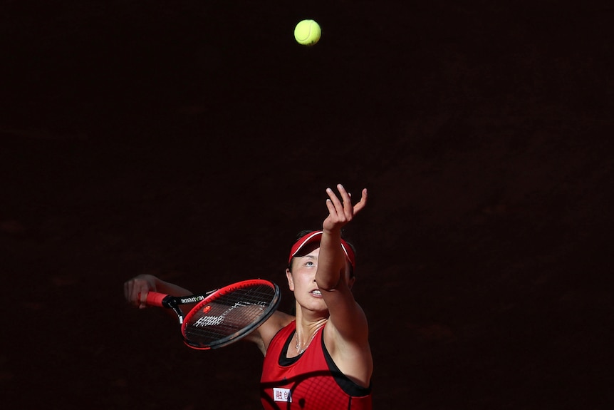 Peng Shuai in a red tennis outfit throws a tennis ball up in the air 