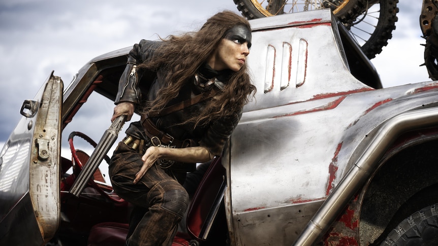 Film still of Furiosa, a woman with the top half of her face painted black, dives out of a vehicle with a gun in hand.