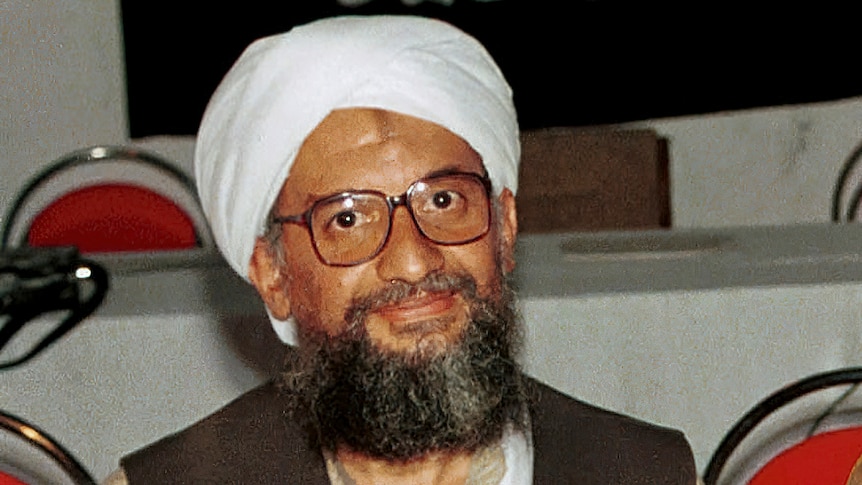 A bearded man wearing a white turban and thick rimmed glasses sits at a table.