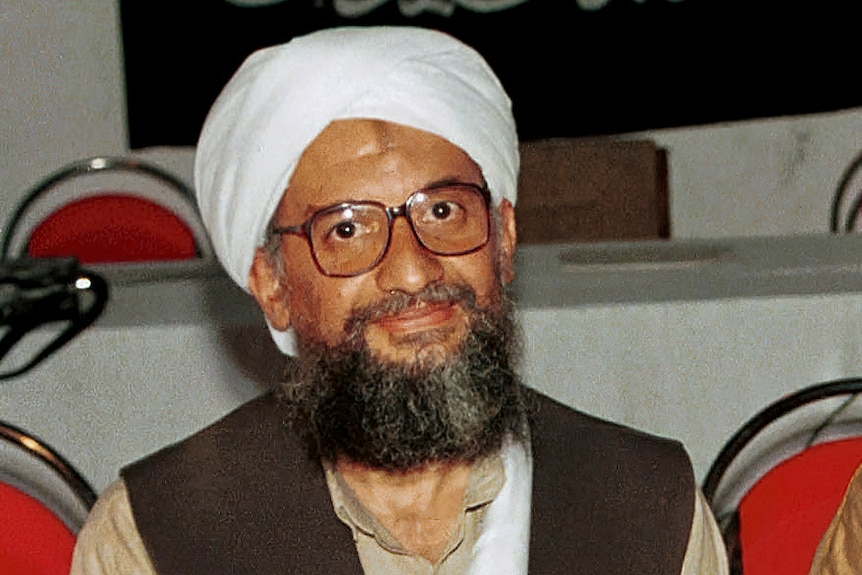 A bearded man wearing a white turban and thick rimmed glasses sits at a table.