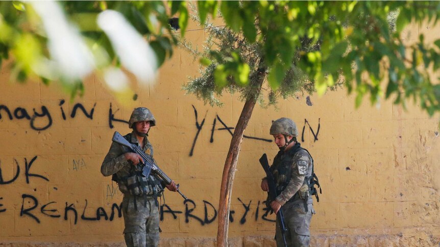 Two soldiers carrying automatic rifles stand under a tree next to a yellow wall.