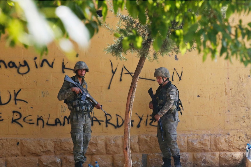 Two soldiers carrying automatic rifles stand under a tree next to a yellow wall.