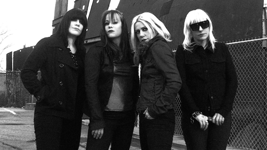 Black and white photo of four-piece rock band L7. All members are women, all wear coats, one wears sunglasses