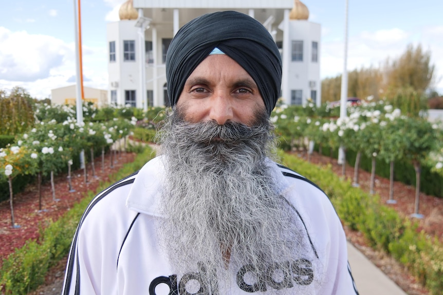 A man wearing a dastar with a long grey and white beard, wearing an adidas jumper and standing in front of a Sikh temple.