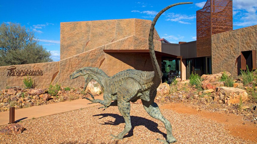 Daytime photograph of the entrance to the Age of Dinosaurs museum, which shows a life size statue of a raptor.