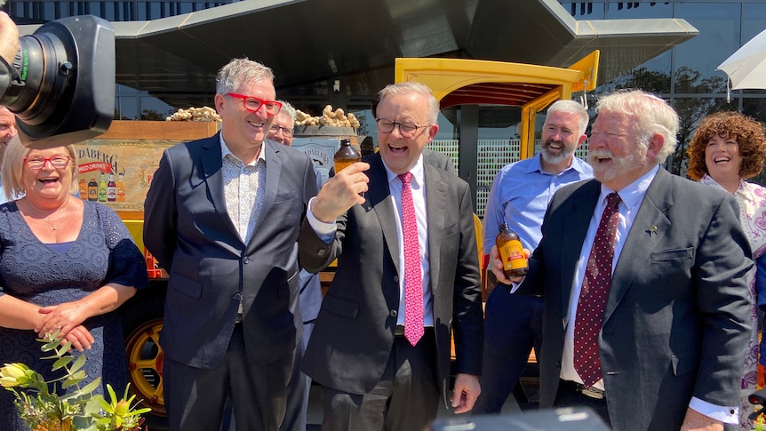 A photo of the PM holding a bottle of ginger beer smiling, surrounded by a group of smiling and laughing people.