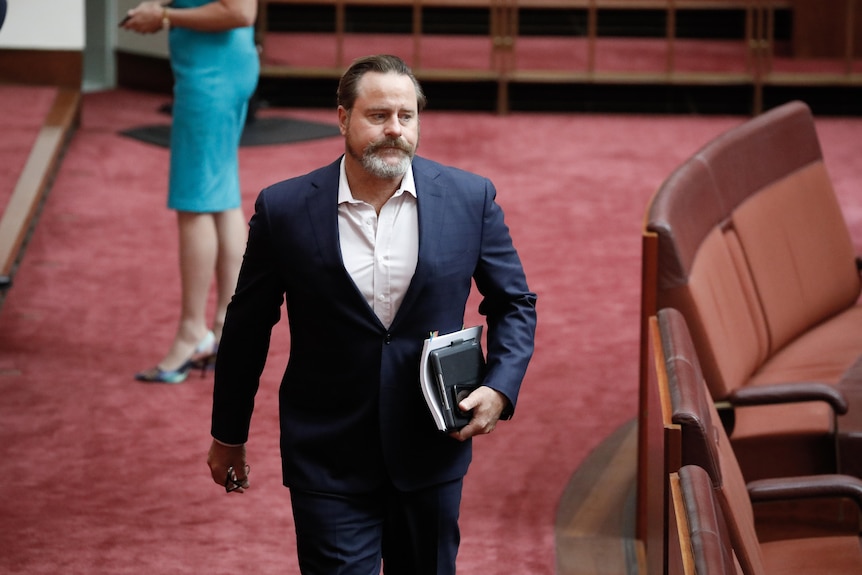 Whish-Wilson is wearing a blue suit, no tie, holding some papers while walking towards camera.