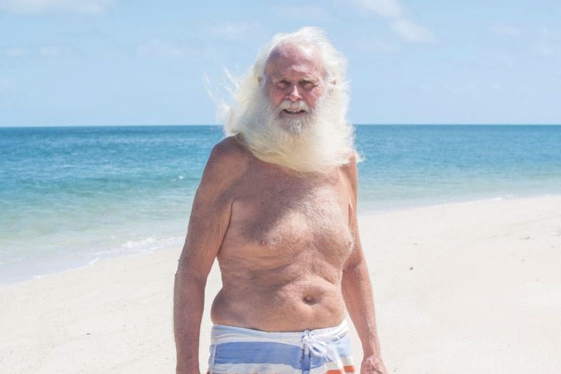 Man with long white hair and beard, wearing boardshorts, stands on white sand with ocean behind him.