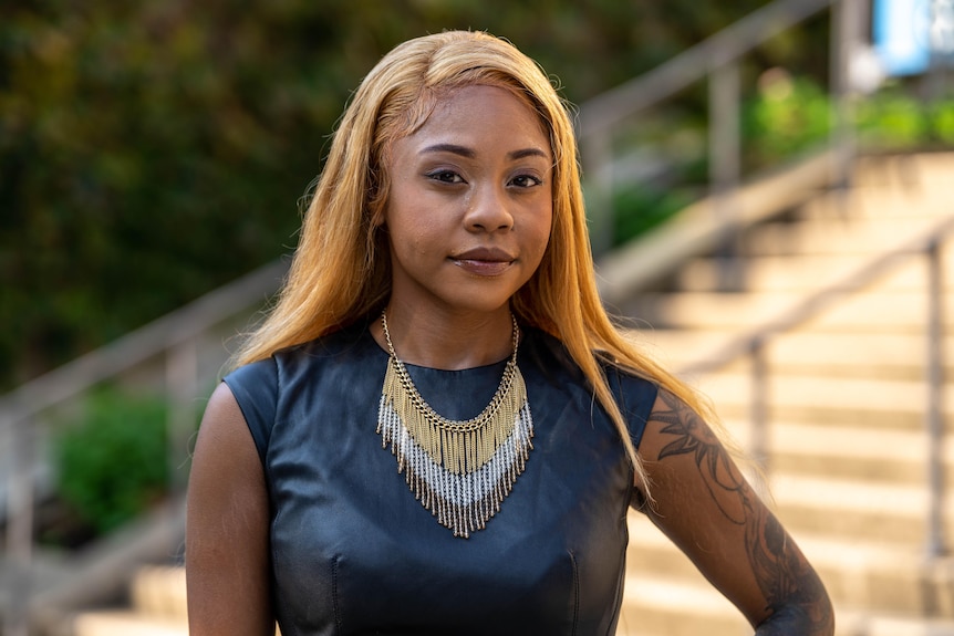 Kyla wears a black leather dress and a large necklace featuring gold and silver. Pictured outside in front of stairs.