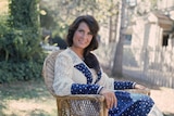 lynn in a cane chair, blue and white lace dress, sits in a sunny backyard