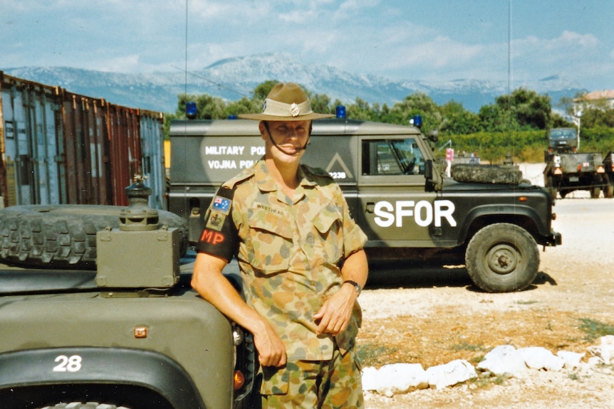 An Australian soldier with am MP armband leaning on a military vehicle with more vehicles in the background.