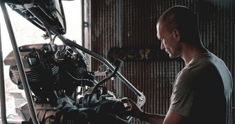 A young man with a shaved head works on an automobile engine in a workshop.