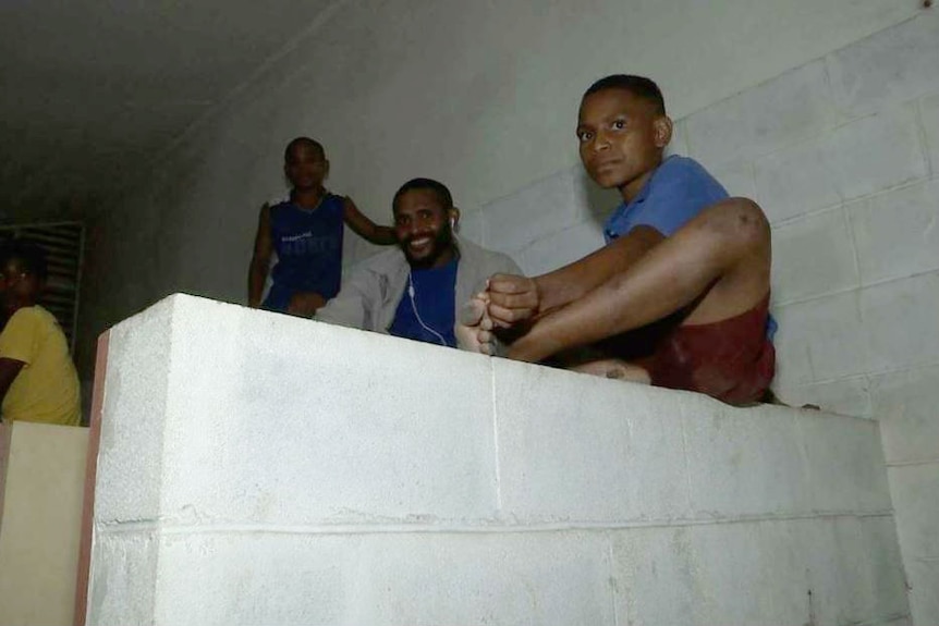 A number of young and older boys are seen sitting in a public toilet cubicle.