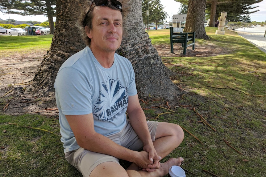 A man sits on grass under a tree looking at the camera.
