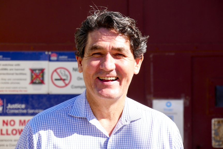 A man with dark hair and a collared shirt on smiles in front of a prison. 