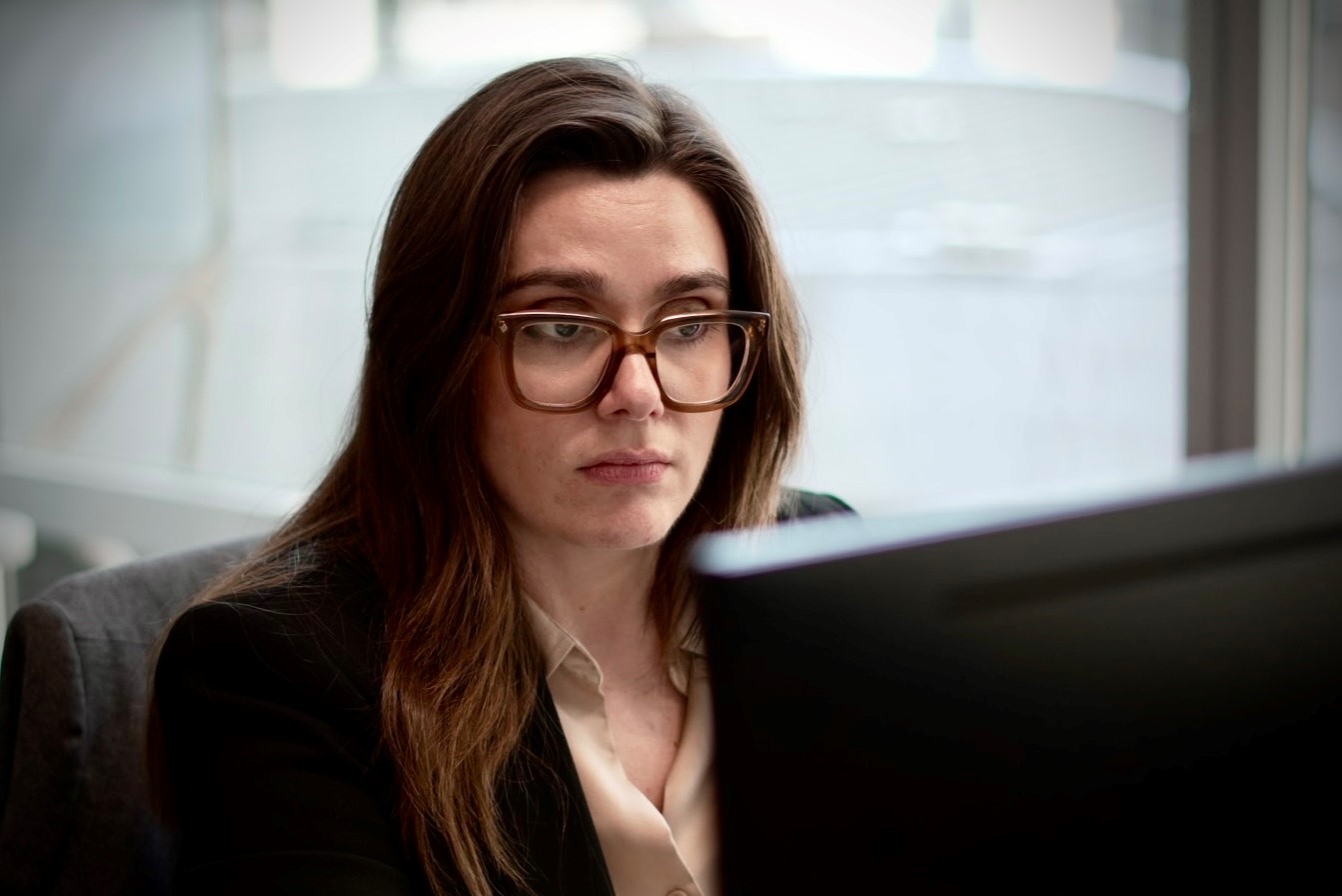 Uma mulher com longos cabelos escuros e óculos marrons de aros grossos está sentada em uma mesa olhando para a tela de um computador.