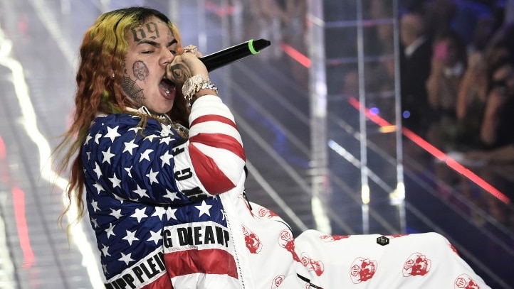 Tekashi 6ix9ine sings into a microphone on stage wearing a US flag jacket