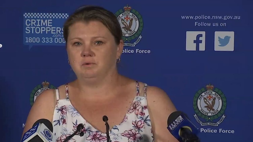 A heavyset woman with brown hair tied back, white blouse, stands before several microphones at a police press conference