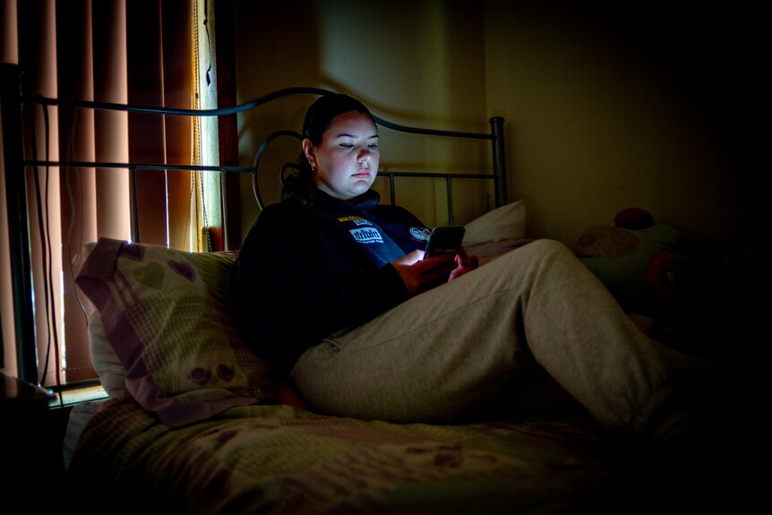 Teenage girl looks at phone in dark bedroom. 